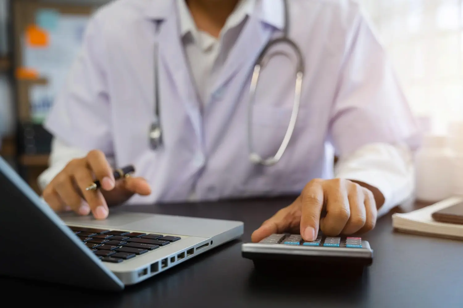Requisitos para a equiparação hospitalar: Foto de um médico utilizando um computador e calculadora para cálculos tributários